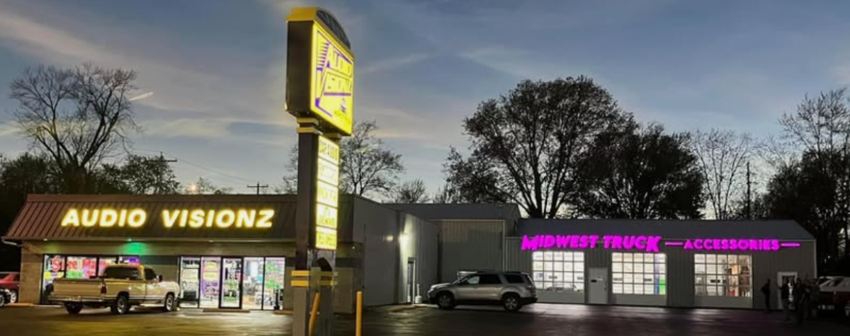 Photo of the Audi Visionz and Midwest Truck Accessories building. Theres a storefront connected to a garage with three garage doors. The Audio Visionz neon sign is in yellow letters. The Midwest Truck Accessories name is in pink neon lights. 