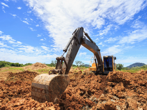 A yellow excavator digs into red dirt. There is a bright, blue sky in the background. 